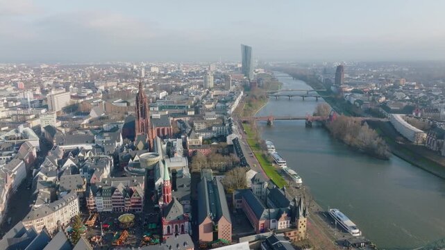 Aerial view of Frankfurt: Main river with bridges and boats, Frankfurt Cathedral and old town on the left, modern skyline in the background under a clear sky.