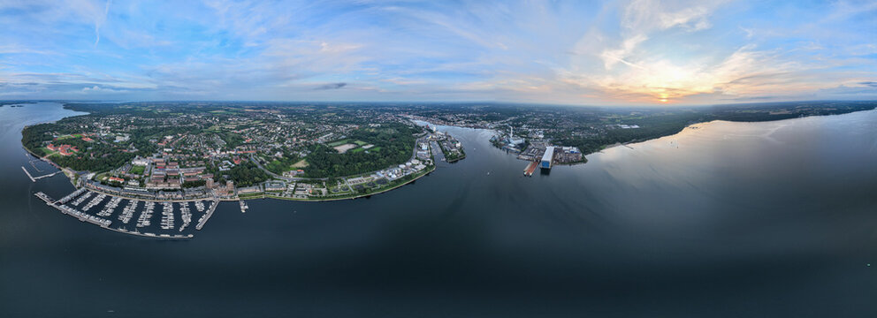 Aerial view of sunlight kissing the horizon over the tranquil waters embracing the city's edge, dotted with boats and industrial zones, Flensburg, Germany.