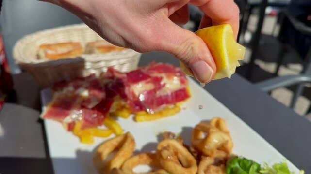 Close-up of a hand squeezing fresh lemon juice over a plate of fried calamari, with serrano ham on the side. Traditional Mediterranean cuisine concept, fresh food and gastronomy. Tapas