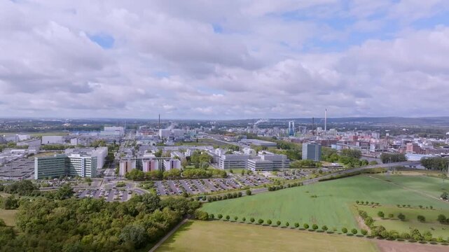 An expansive aerial view of a modern industrial park or manufacturing facility, surrounded by large green areas and parking lots. In the distance, factory halls, chimneys, and office buildings can be 