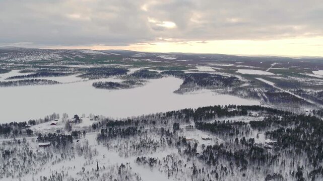 Impressive aerial shot of a vast, wintry lake district in Finland. Dense, snow-covered coniferous forests surround the frozen white waters under an overcast sky.