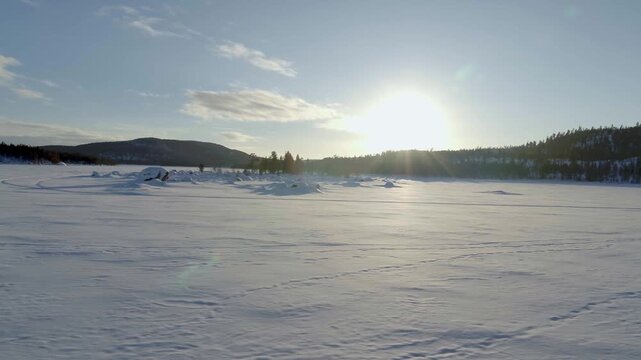The low winter sun reflects off a vast, pristine expanse of snow in Finland. In the background, dark forests and gentle hills stretch out under a clear, bright sky.