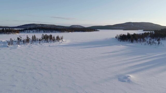 A vast, deeply snow-covered winter landscape in Finland featuring a completely frozen lake, surrounded by dense coniferous forests and gentle hills in the soft light of the low sun.