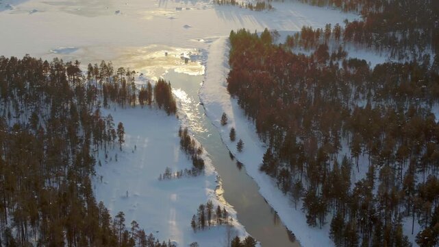 An impressive aerial shot from Finland, showing a narrow, ice-free river winding its way through deeply snow-covered, dense coniferous forests in atmospheric lighting.