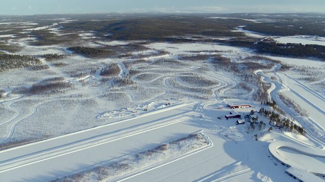 A vast aerial view of a winter testing track or facility in Finland. White tracks and paths wind through the snowy plain, complemented by red wooden houses under the bright sunlight.