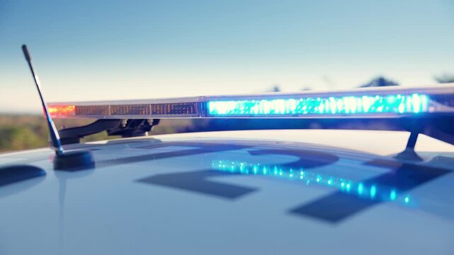 Close-up of police car light bar with red and blue emergency lights flashing brightly on vehicle roof. Antenna visible against clear blue sky with blurred trees in background during daytime patrol.