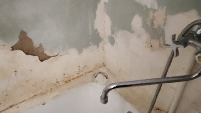 Person approaching dirty shabby bathroom with mold and peeling plaster, turning on water tap, first person view