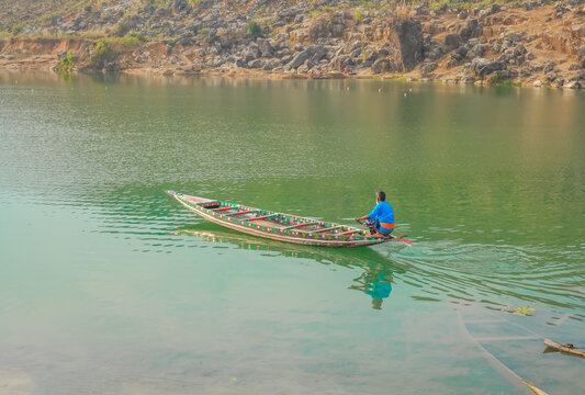 Sunamganj, Bangladesh - 26 January 2019: View of a lone boatman gently rowing across the tranquil, emerald waters, framed by the rugged, rocky terrain of the quarry hills.