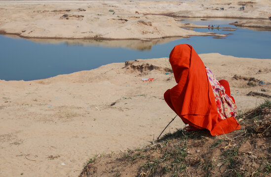 Sunamganj, Bangladesh - 26 January 2019: View of a woman in vibrant orange attire crouches pensively by the sandy riverbank, her silhouette contrasting against the shimmering water.
