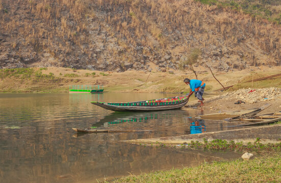 Sunamganj, Bangladesh - 26 January 2019: View of a lone figure tending to a vibrant, narrow boat on the tranquil, reflective waters against a backdrop of rugged, earthy cliffs.