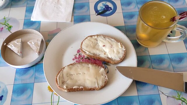 Two cheese sandwiches on plate with knife, glass mug of tea, wrapped cheese triangles, breakfast ready on kitchen table
