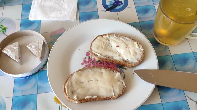 Two cheese sandwiches on plate with knife, glass mug of tea, wrapped cheese triangles, breakfast ready on kitchen table