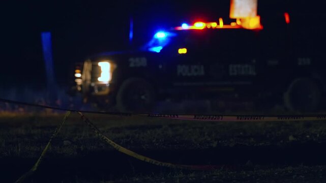 State police truck with red and blue flashing lights illuminates a night crime scene. Yellow caution tape stretches across grass in foreground with emergency vehicle in background.