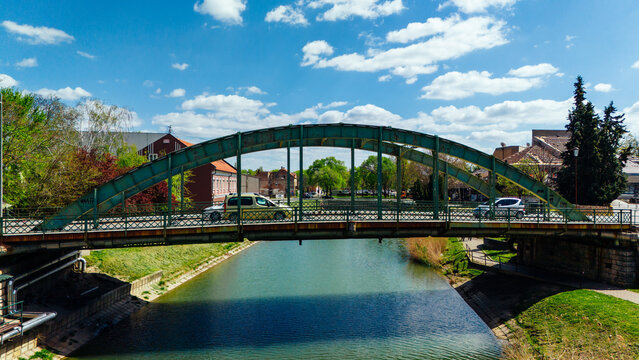 Aerial view of a curved green bridge over calm waters reflecting the sky, with cars crossing, set against the backdrop of the city, Zrenjanin, Vojvodina, Serbia.