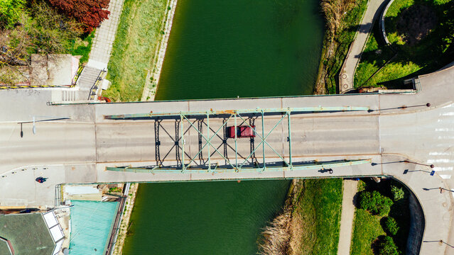 Aerial view of a red car crossing a bridge over a green canal, with concrete banks and hints of spring foliage, Zrenjanin, Vojvodina, Serbia.