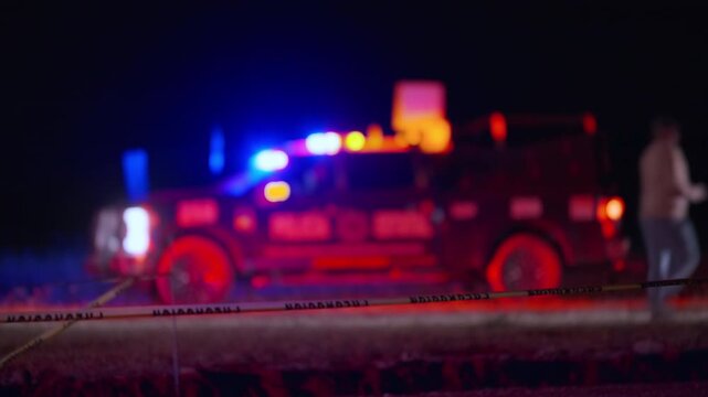 Defocused fire truck with red and blue emergency lights flashing behind yellow caution tape at night scene. Bokeh effect creates dramatic atmosphere at accident site with glowing orbs.