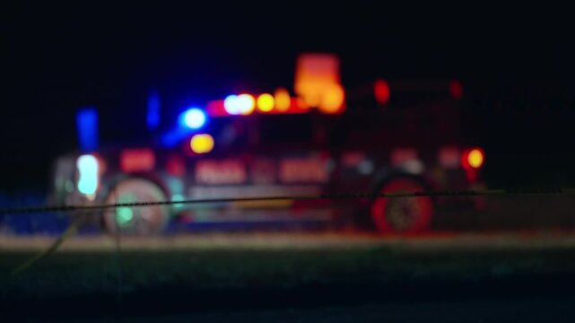 Defocused fire truck with flashing red and blue emergency lights behind yellow caution tape at night scene. Bokeh effect creates dramatic atmosphere at accident site with colorful glow.