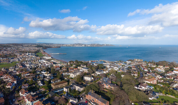 Aerial view of the vibrant coastal town where red-roofed buildings meet the serene blue sea, framed by the distant hills and a bright sky over Paignton, England, United Kingdom.