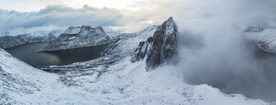 Aerial panoramic view of craggy peaks pierce through a blanket of fresh snow, their dark rock faces contrasting sharply against the pristine white landscape. Segla, Senja, Troms, Norway.