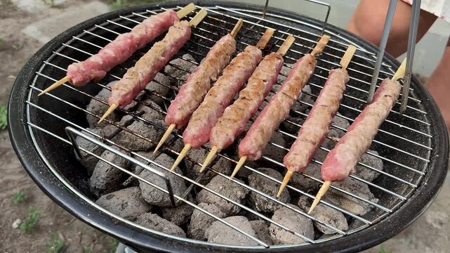 Skewers of minced meat cook over charcoal on a round grill, outdoor barbecue scene with glowing coals, warm casual mood for summer gatherings