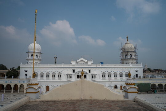 View of the serene white Gurdwara bathed in sunlight, its domes reaching towards the tranquil blue sky, a golden flag standing tall, Nankana Sahib, Punjab, Pakistan.