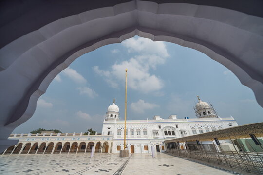 View of the crisp white Gurdwara framed by an ornate arch, gleaming under a vast sky, a testament to serenity and architectural beauty, Nankana Sahib, Punjab, Pakistan.