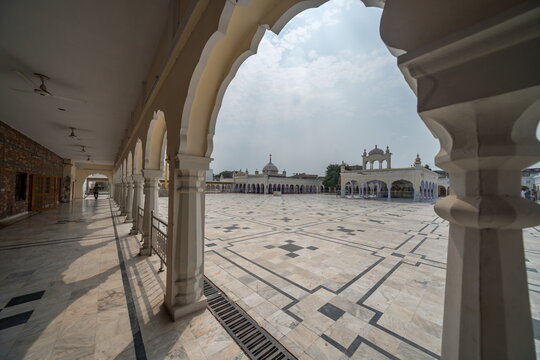 View of the expansive courtyard with geometric patterned marble flooring, framed by ornate arches and buildings under a cloudy sky, Nankana Sahib, Punjab, Pakistan.
