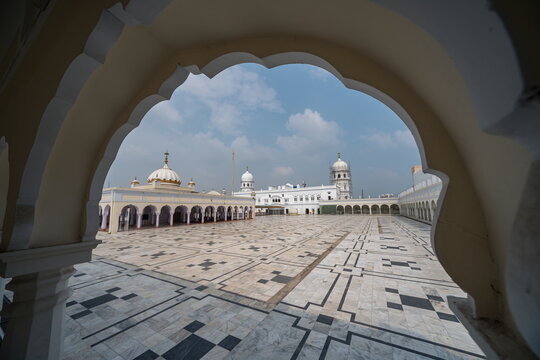 View of the expansive marble courtyard with intricate geometric patterns framed by ornate arches, leading to pristine white buildings, Nankana Sahib, Punjab, Pakistan.