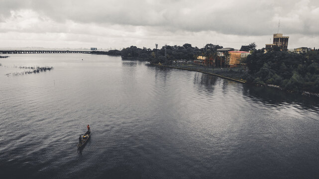 Aerial view of a canoe cutting through the still, dark waters towards the distant, hazy horizon near the buildings of Oworonshoki, Lagos, Nigeria.