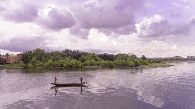 Aerial view of a lone canoe cuts through the still water, reflecting the cloudy sky, with lush greenery lining the distant shore, Oworonshoki, Lagos, Nigeria.