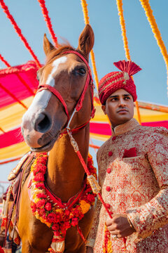 Modern indian groom standing beside horse during traditional baraat wedding procession, colorful festive decorations and bright daylight, cultural celebration atmosphere, background with blank space