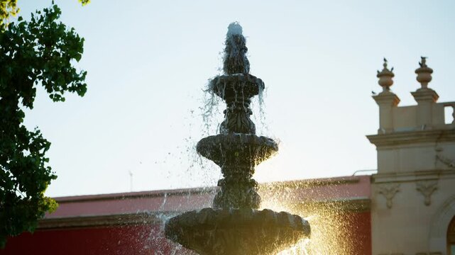 Elegant multi-tiered stone fountain cascades water droplets illuminated by warm golden hour sunlight. Historic colonial architecture with decorative finials and red wall creates scenic backdrop.