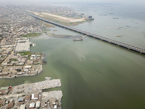 Aerial view of a long bridge stretching towards the horizon over murky waters, contrasting with the chaotic sprawl of buildings and waste, Oworonshoki, Lagos, Nigeria.