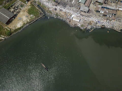 Aerial view of the riverbank choked with refuse next to simple buildings, a stark contrast to the flowing water and lone boat, Oworonshoki, Lagos, Nigeria.
