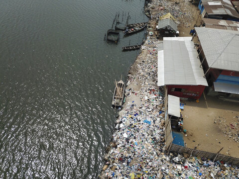 Aerial view of the shoreline's stark contrast between the dark water and the overwhelming piles of colorful trash near buildings, Oworonshoki, Lagos, Nigeria.