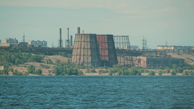 A sprawling industrial complex, featuring large cooling towers and smokestacks, stands on the shore of a wide river. Muted colors and hazy light create a somber, atmospheric mood. Concept for energy