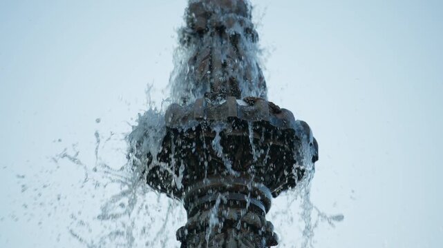 Close-up view of an ornate tiered fountain with water splashing and cascading over decorative bronze sculpture. Golden sunlight illuminates droplets against a pale sky background.