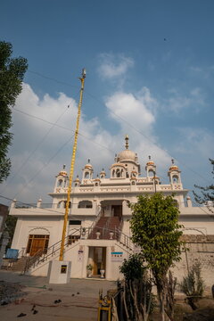 View of a tall, golden Nishan Sahib flag standing proudly before a pristine white Sikh temple under a vast blue sky, Nankana Sahib, Punjab, Pakistan.