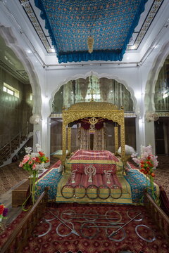 View of a golden palanquin with ornate carvings rests under a blue floral ceiling, bathed in soft light, in the serene sanctuary, Nankana Sahib, Punjab, Pakistan.