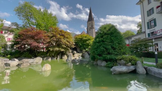 Static shot of serene Japanese garden with green koi pond, rocks, lush trees, and church steeple under partly cloudy sky in Interlaken, Switzerland.