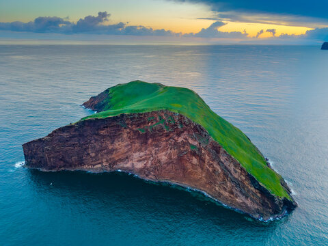 Aerial view of a striking island with vibrant green vegetation contrasting against its rugged, reddish-brown cliffs surrounded by the deep blue sea, Terceira, Portugal.