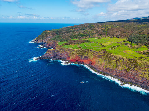 Aerial view of Ilheus das Cabras' rugged coastline where deep blue sea meets vibrant green fields and striking red cliffs, Terceira, Azores, Portugal.