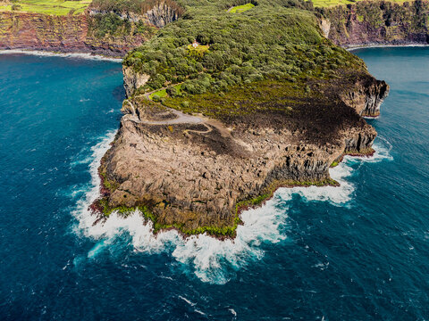 Aerial view of the dramatic Ilheus das Cabras islet, with its rugged, dark volcanic rocks meeting the turquoise sea, contrasted by the lush green vegetation, Terceira, Azores, Portugal.