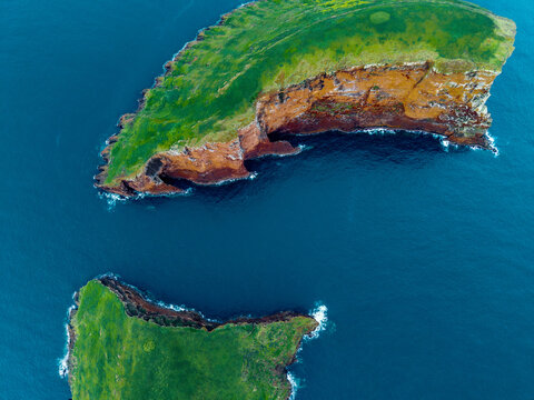 Aerial view of Ilheus das Cabras islands showcasing the vibrant green vegetation contrasting with the reddish-brown cliffs against the deep blue ocean, Terceira, Azores, Portugal.