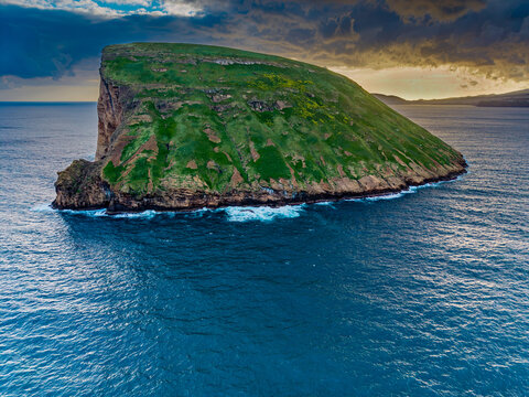 Aerial view of Ilheus das Cabras island, a verdant jewel rising from the turquoise waters, fringed by white waves under a dramatic sky, Terceira, Azores, Portugal.