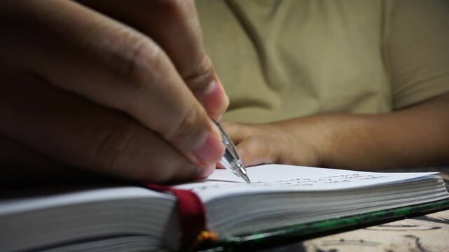 A male finishes writing on a page before closing a green notebook in a dark room, capturing the end of a journaling session with a calm aesthetic.