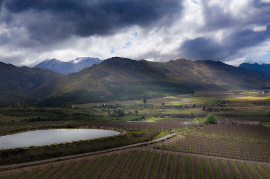 Aerial view of dramatic sunlight highlighting the vineyards and dam against the backdrop of rugged mountains under a stormy sky., Cape Town, Western Cape, South Africa.