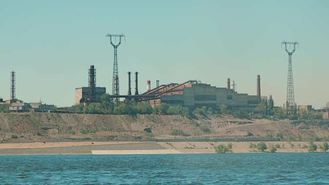 A sprawling industrial complex with smokestacks sits on a high riverbank. Power lines stretch across a clear sky. The scene, with its muted tones and teal water, evokes themes of industry and energy
