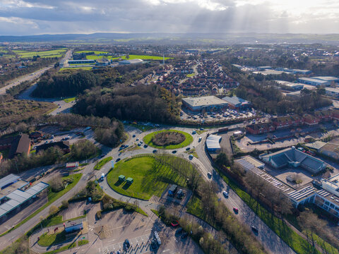 Aerial view of a roundabout bustling with cars near a shopping center, framed by lush greenery and distant fields, Exeter, England, United Kingdom.