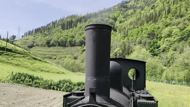Vista en primer plano de la chimenea de una locomotora de vapor antigua expulsando humo denso mientras circula por una v&iacute;a rural bajo un cielo despejado.